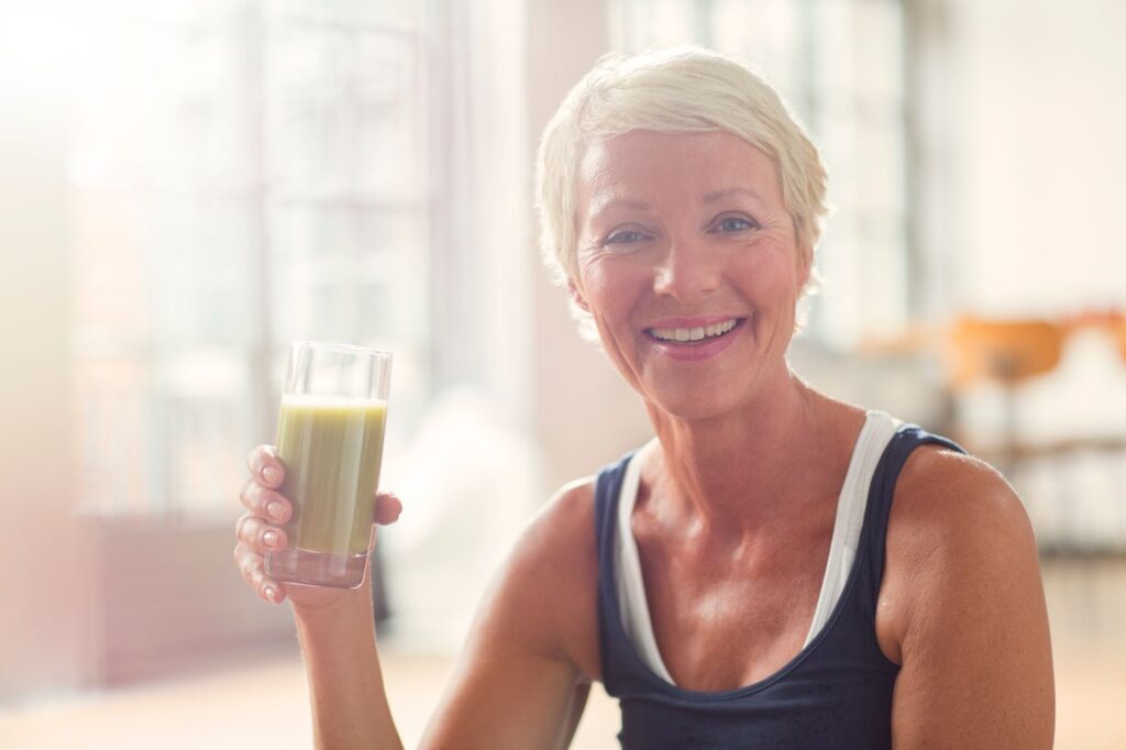 An older woman smiling and holding a home-made meal replacement smoothie in her right hand.